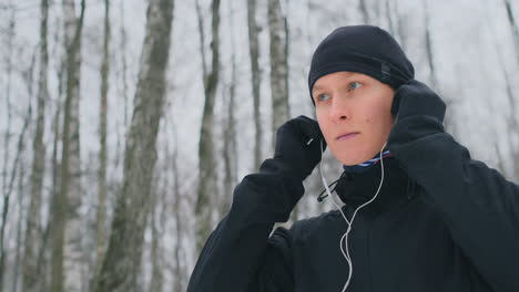 A-young-man-on-a-morning-jog-holds-headphones-in-his-hands-and-inserts-it-into-his-ears-before-running-in-the-park.-Winter-run.-Use-modern-gadgets-and-training-applications.-Listen-to-music-while-training-while-running